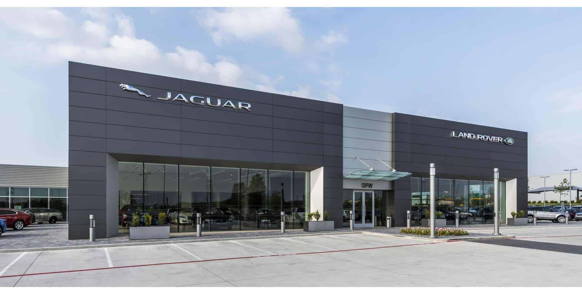 Modern Jaguar Land Rover dealership building with large glass windows, prominent brand logos, and a mostly empty parking lot in front under a partly cloudy sky.