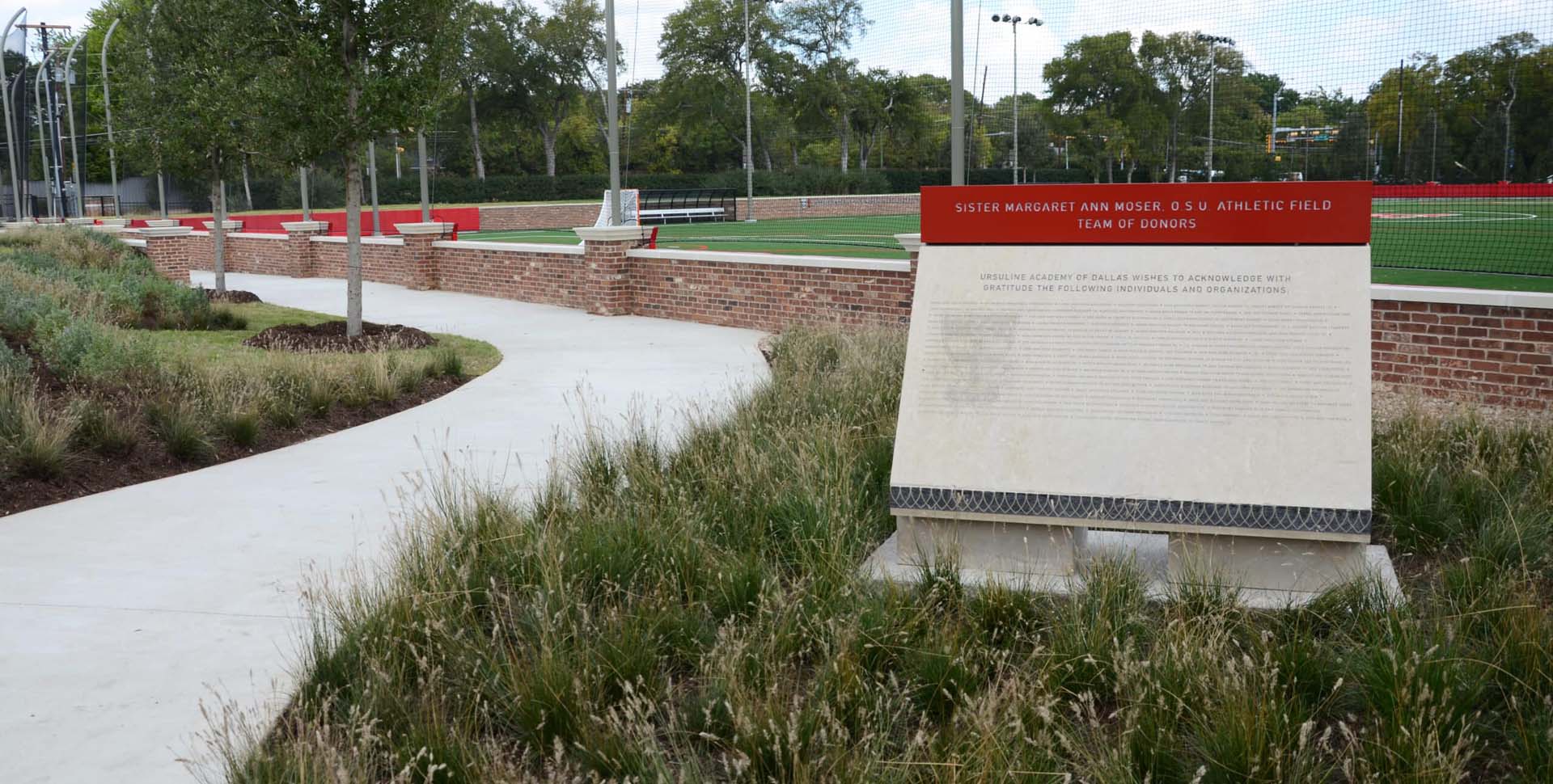 A curved sidewalk runs alongside a brick wall and a plaque for the Sister Margaret Ann Woker S.U. Athletic Field at a sports complex, bordered by grass and trees.