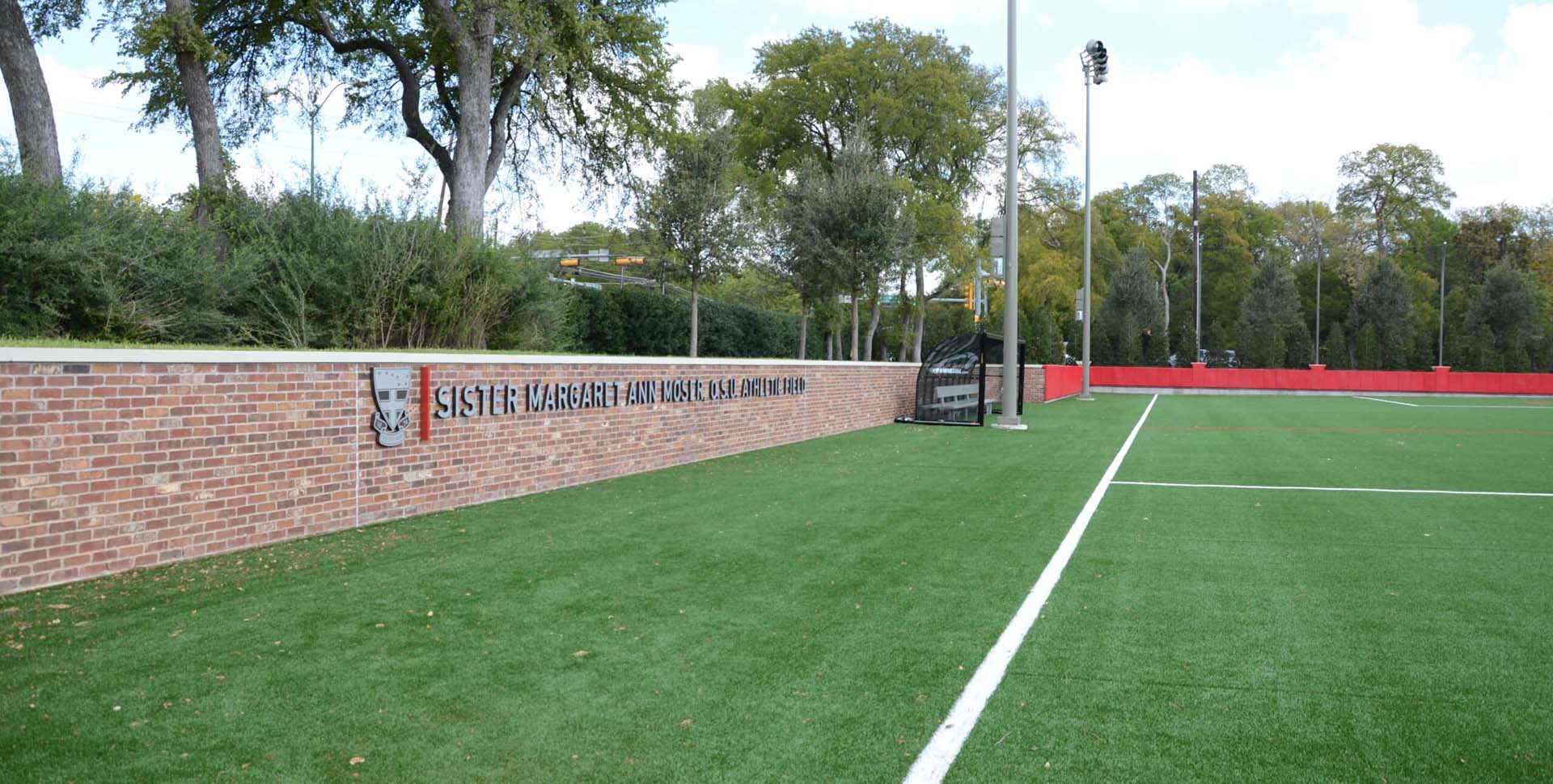 A synthetic turf sports field with white boundary lines, a brick retaining wall, and signage reading "Sister Margaret Ann Moser, CSB, Athletic Field.