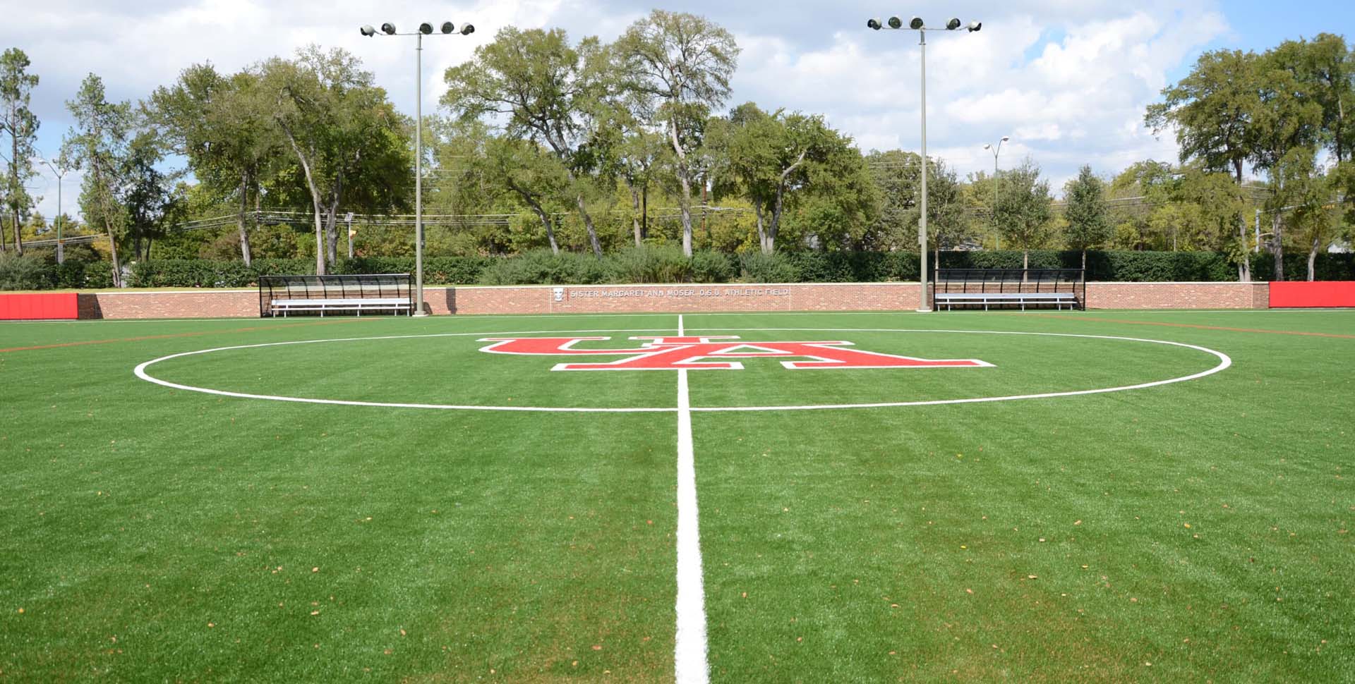 A soccer field with artificial turf, white field markings, a large red logo at the center, benches along the far sideline, and trees in the background.