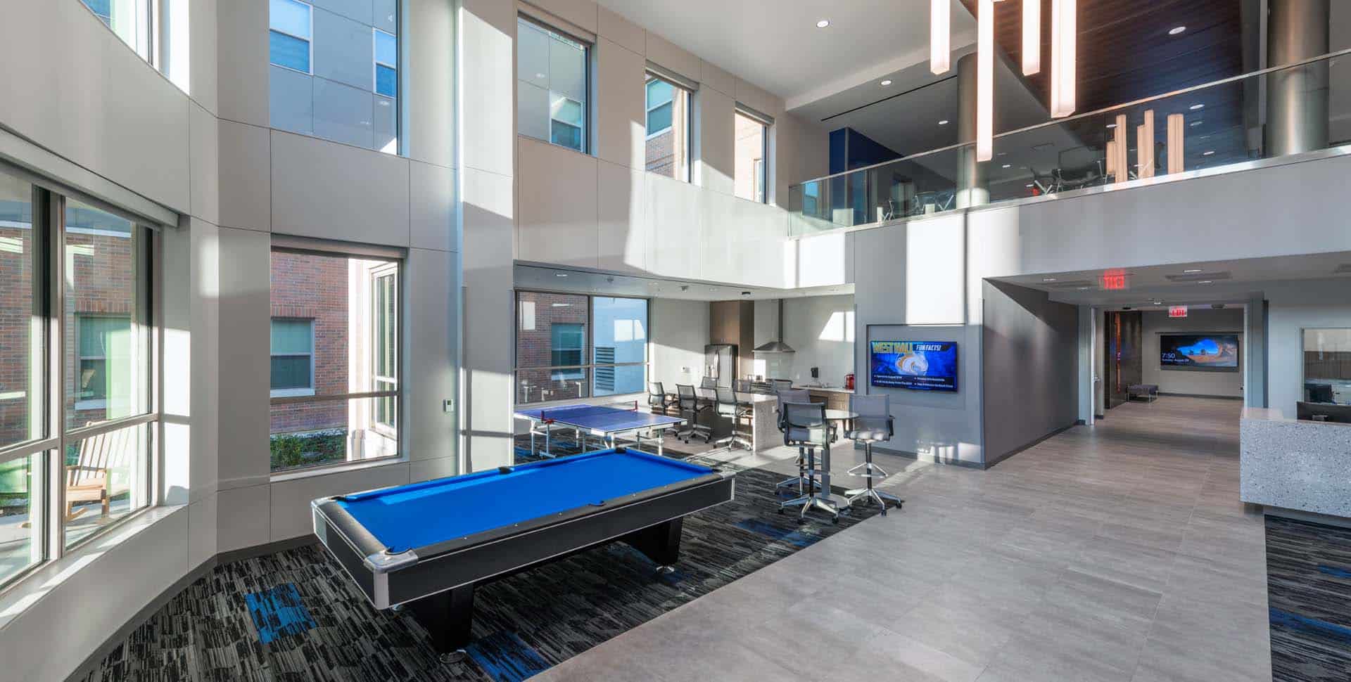 A modern lounge area with a blue pool table, ping pong table, tall chairs, large windows, and a TV, located in a bright, open building interior.