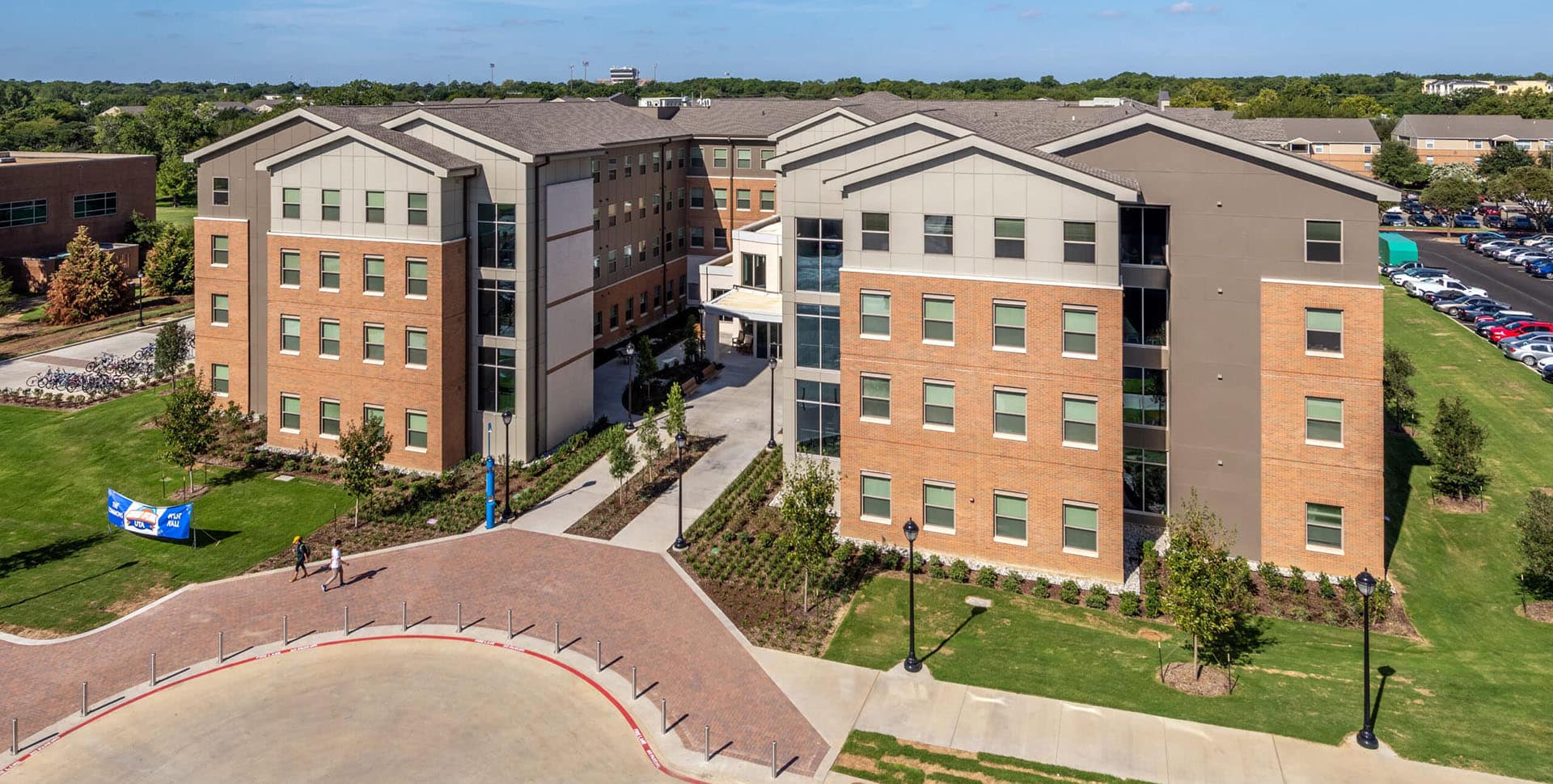 A large, four-story brick and gray building with landscaped grounds and a circular driveway, likely a residential or campus facility, under a clear blue sky.