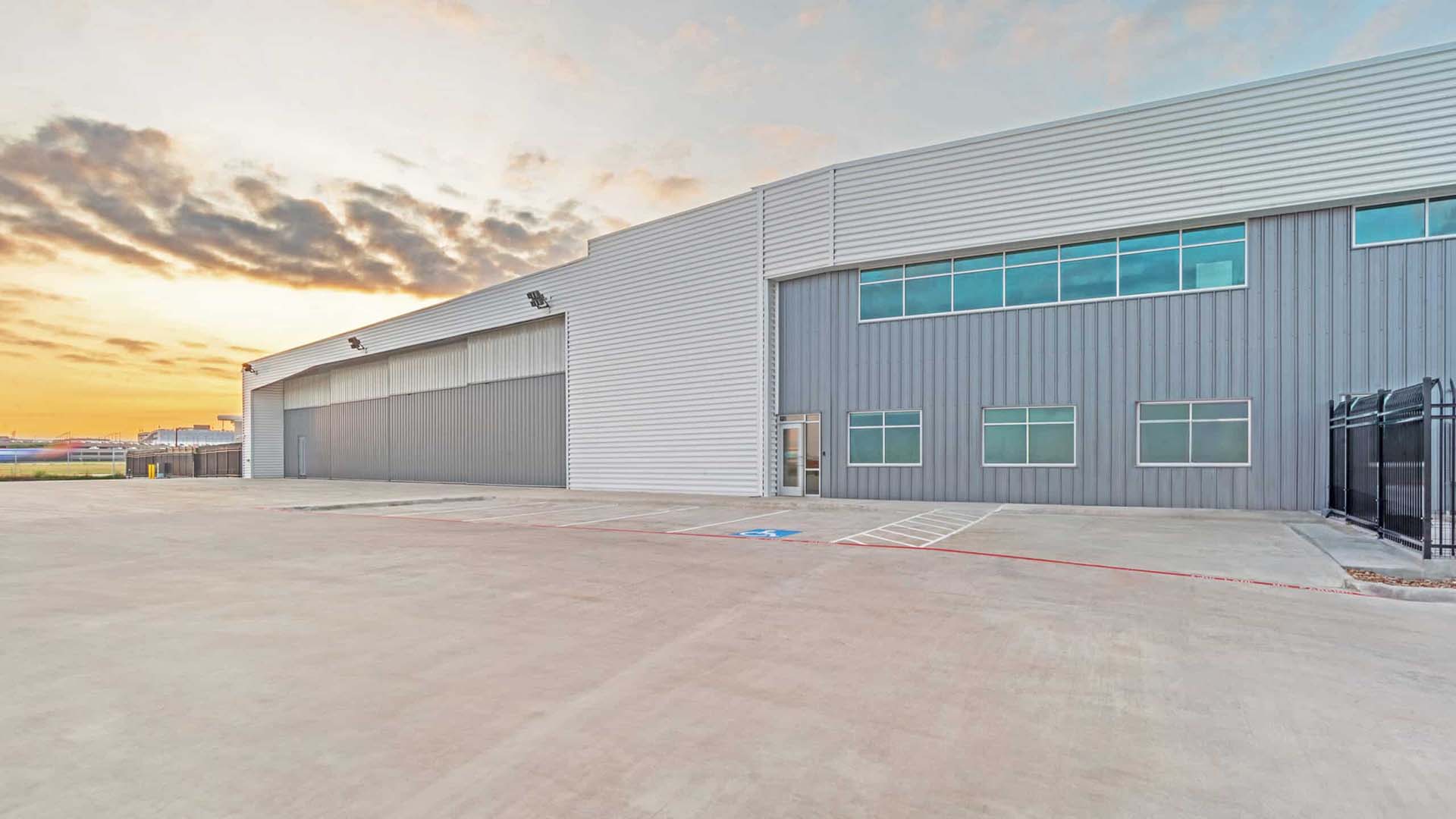 Large industrial warehouse building with metal siding, multiple windows, and a wide driveway at sunset.