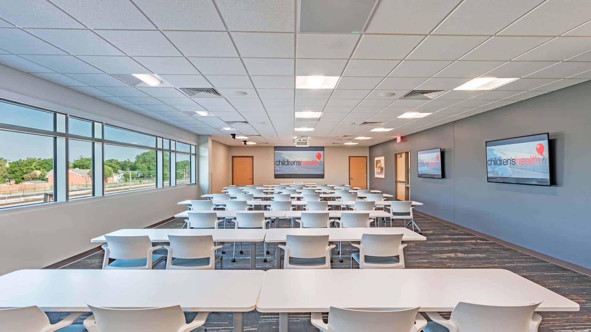 Modern classroom with white tables and chairs, large windows, two wall-mounted screens displaying "children's health," and carpeted flooring. Room is empty and brightly lit.