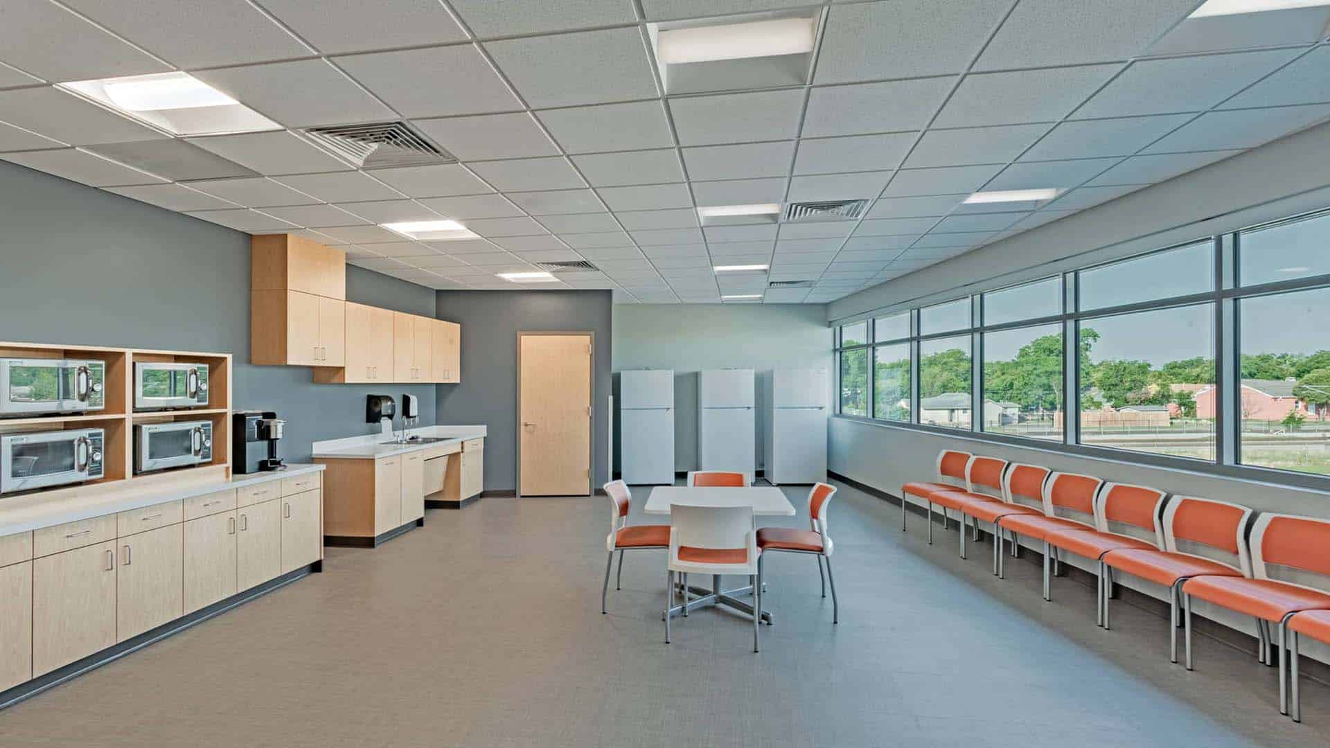 Modern break room with a row of orange chairs, a central table with four chairs, large windows, refrigerators, microwaves, and coffee machines along the wall.