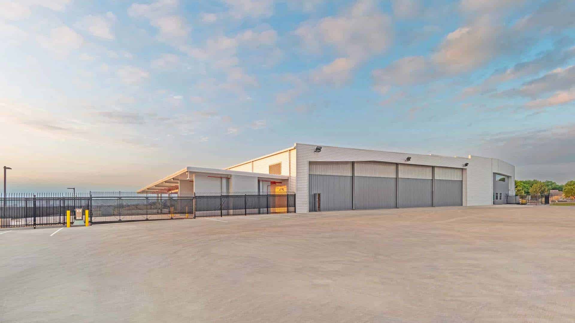 Large, modern industrial warehouse with metal siding and closed roll-up doors, surrounded by a paved lot and a black security fence under a partly cloudy sky.