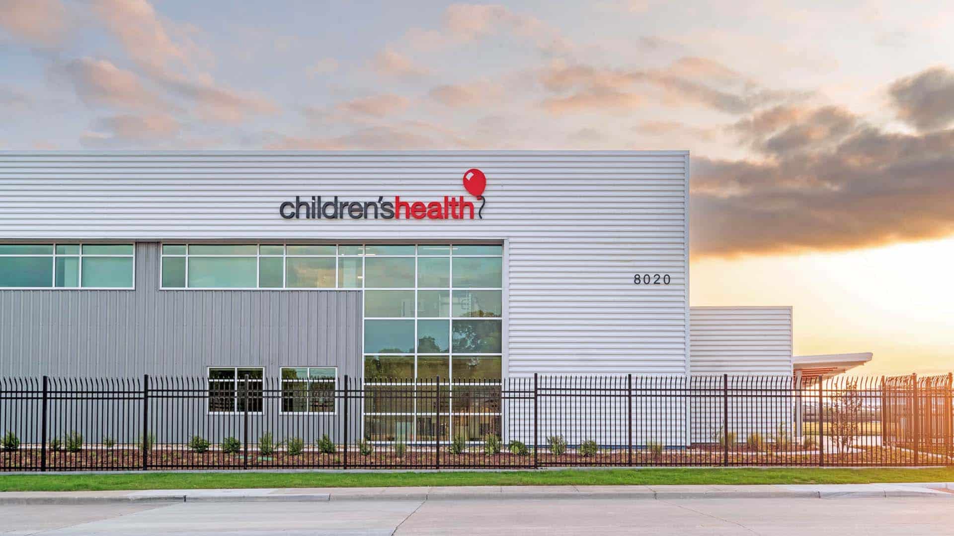 Exterior view of a modern building labeled "children's health" with large windows, a metal fence, and the address number 8020 at sunset.