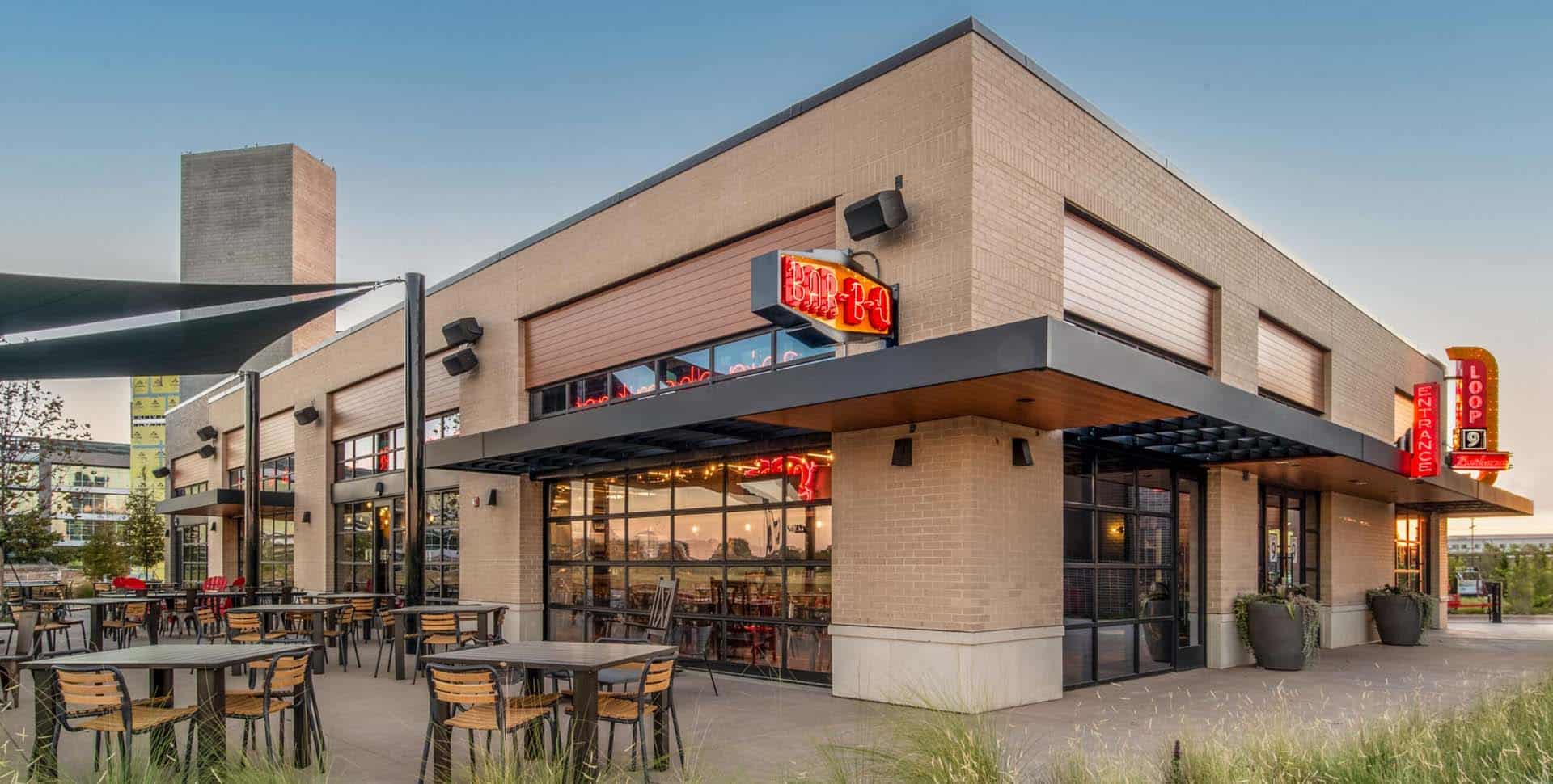 A modern restaurant with outdoor seating, large windows, and neon signs on the exterior, photographed during daylight.