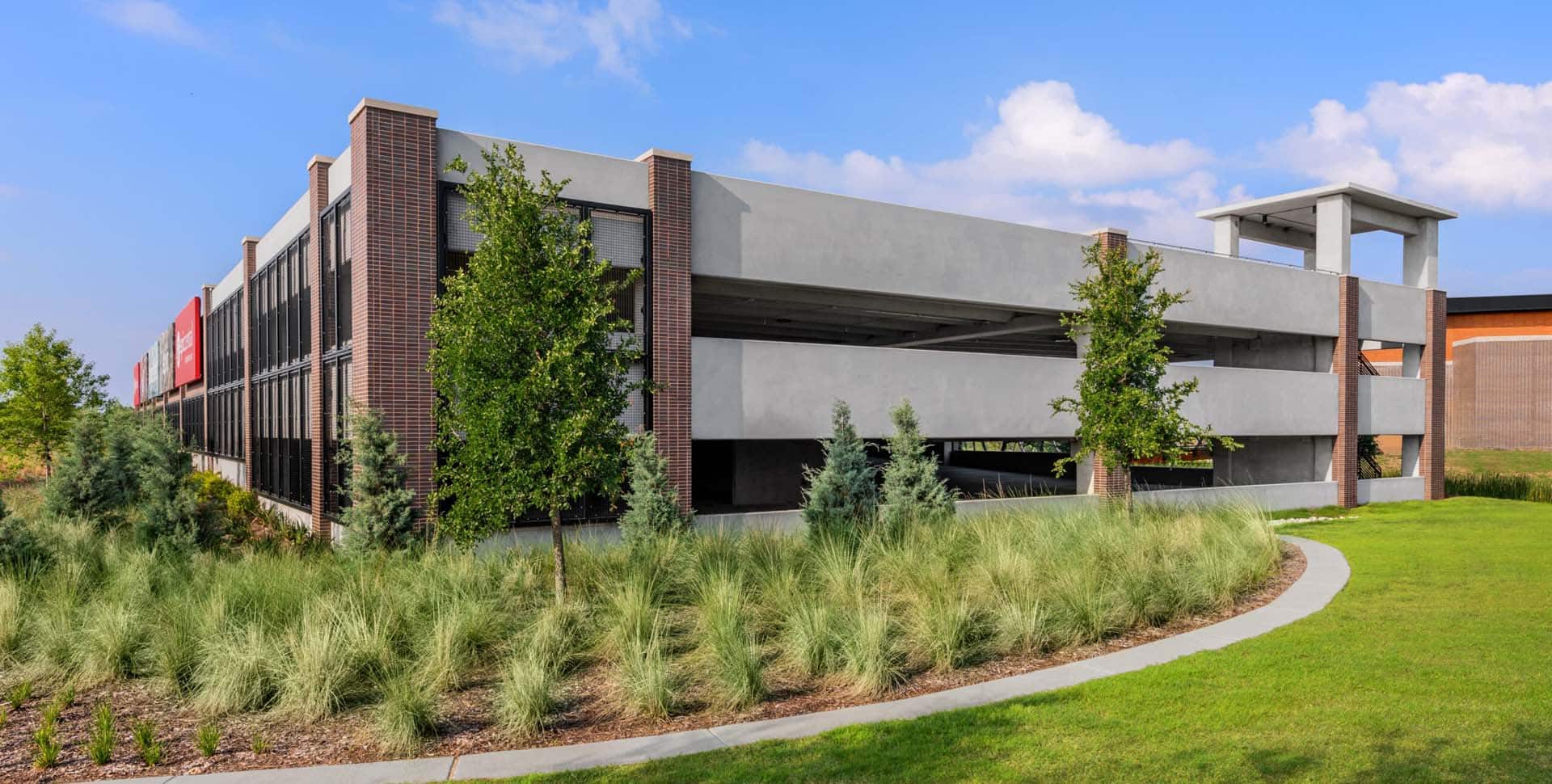 A modern multi-level parking garage with brick and concrete exterior, surrounded by green landscaping and trees under a partly cloudy sky.