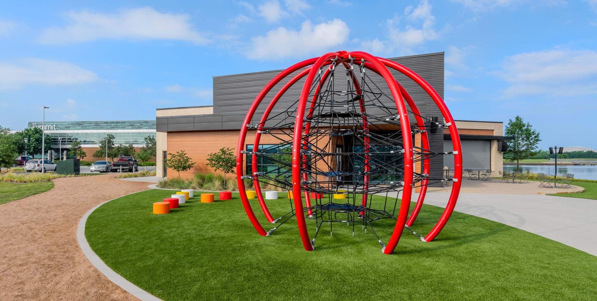 A modern playground structure with red arches and netting stands on artificial grass near a building, with colorful stools and a path nearby.