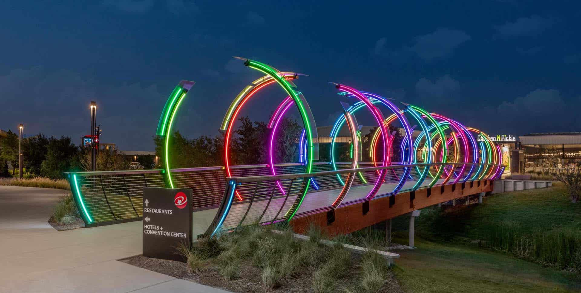 A pedestrian bridge with circular neon light arches in various colors, illuminated at night near a sign for restaurants and a convention center.