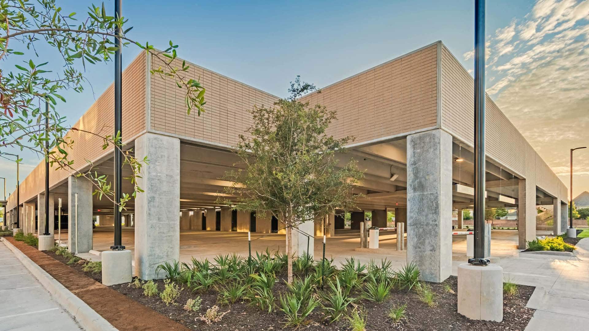 A two-story parking garage with brick and concrete exterior, surrounded by landscaped plants and trees, photographed at sunset.