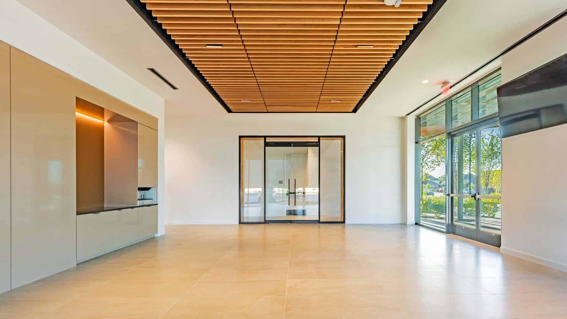Modern, empty office lobby with large glass doors, beige tile flooring, wooden slat ceiling feature, and floor-to-ceiling windows letting in natural light.