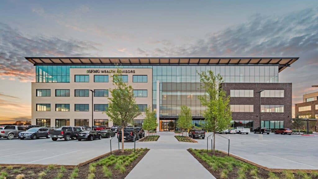 Modern office building with large windows, labeled “Aspire Wealth Advisors,” surrounded by a parking lot and landscaped greenery at sunset.