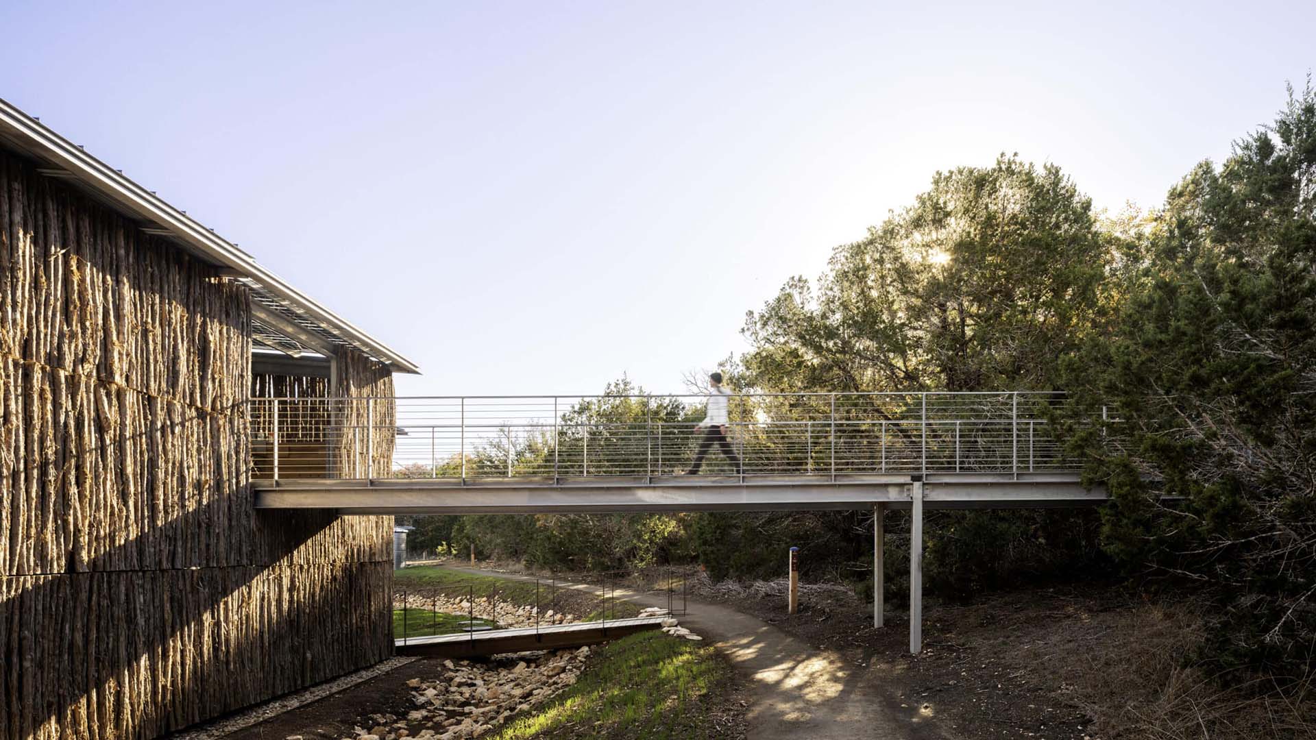 A person walks across a metal pedestrian bridge connecting a wooden building to a wooded area on a sunny day.