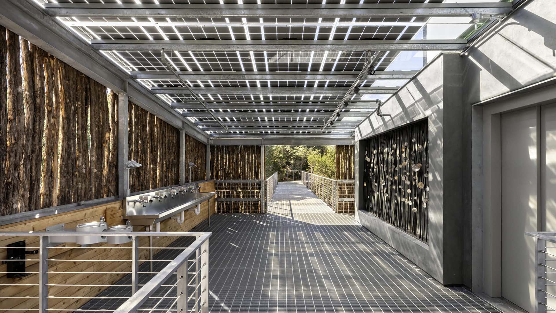 A covered walkway with a grated metal floor and glass roof panels, featuring wooden slat walls and a stainless steel sink along one side.