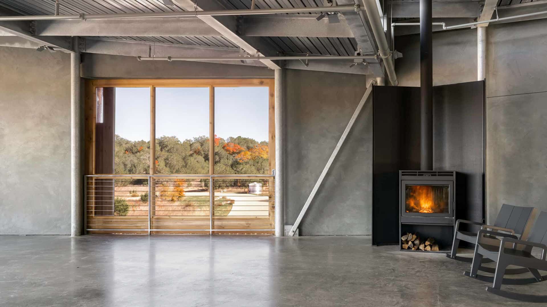 A minimalist room with concrete floors, a black wood stove, a metal chair, and large windows overlooking trees with autumn foliage.