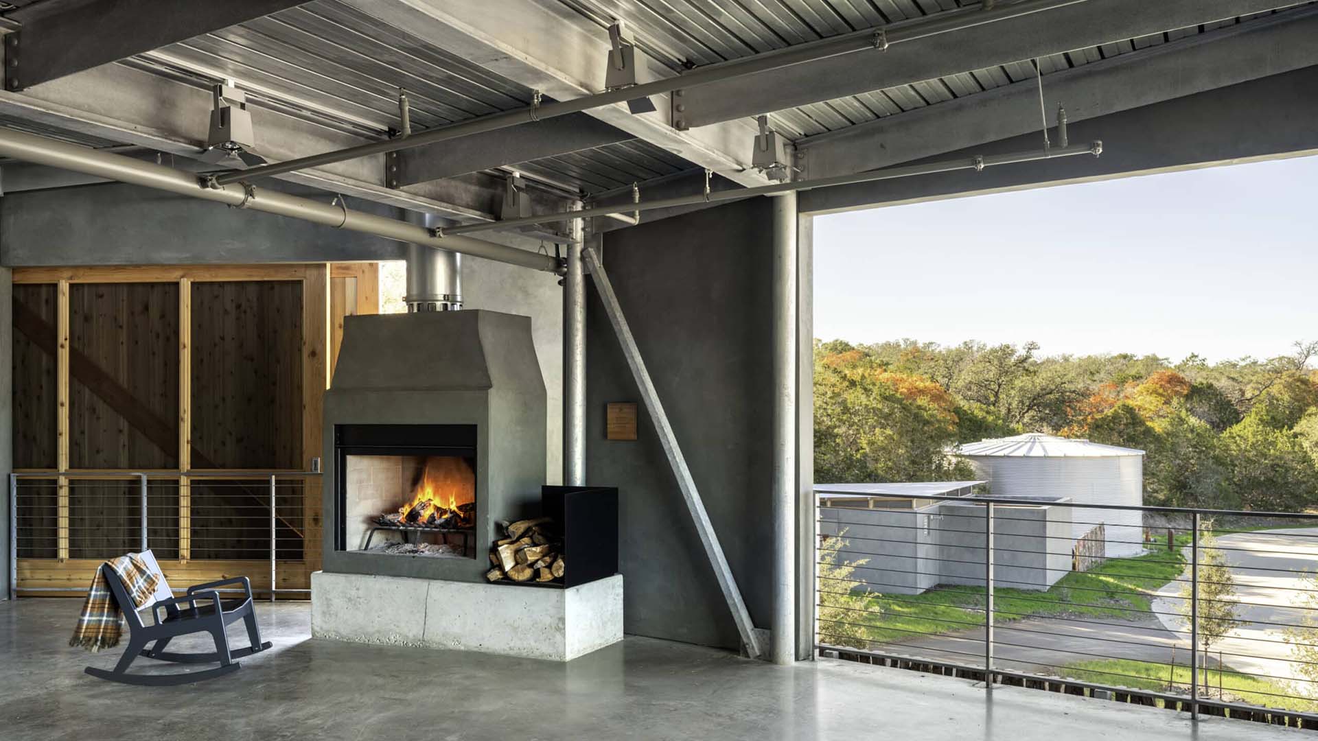A modern industrial-style room features a concrete fireplace, a rocking chair, and an outdoor view of trees and a metal silo building.