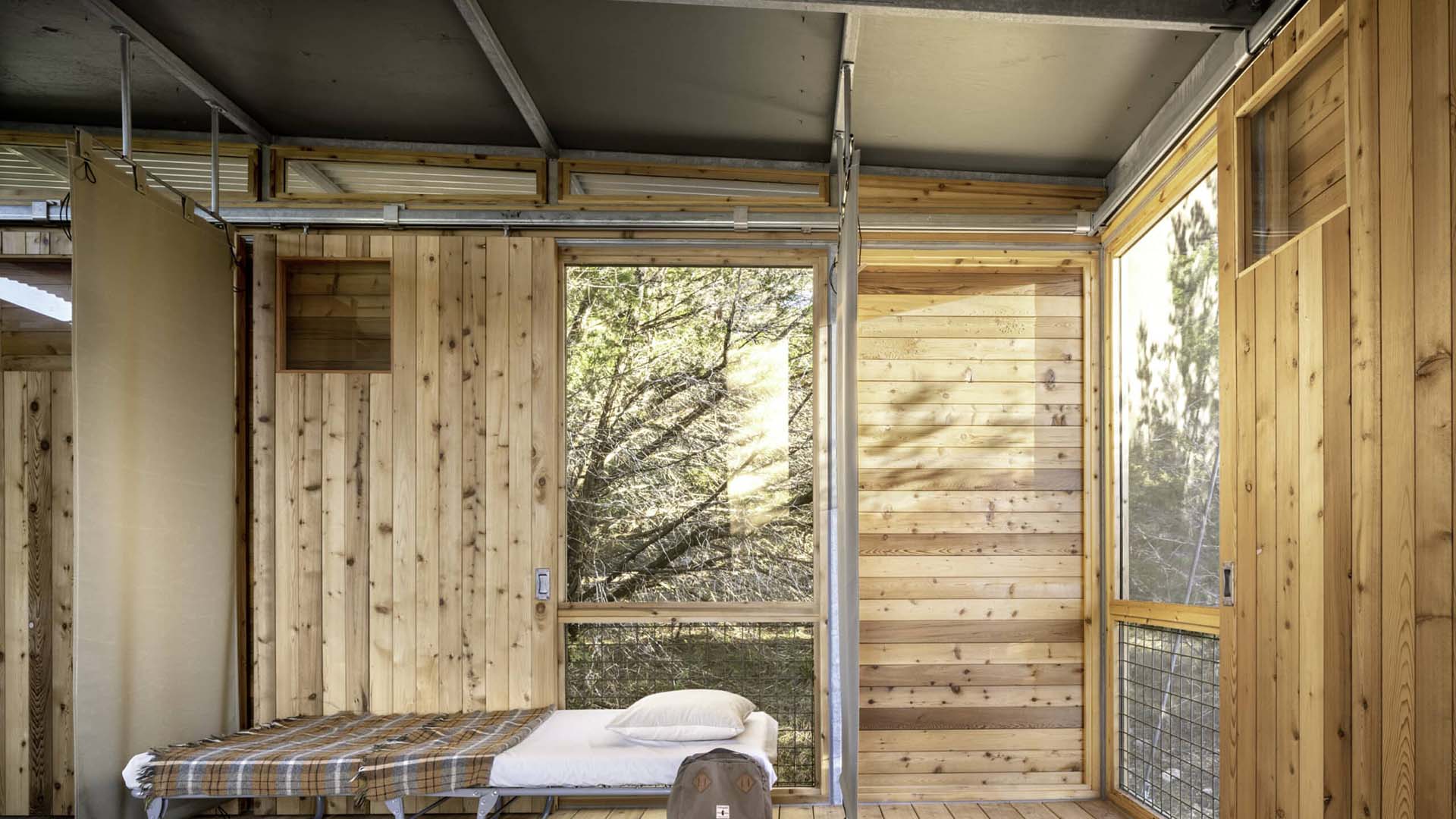 A small room with wooden walls, a metal ceiling, a single bed with a plaid blanket, a pillow, and large windows showing trees outside.