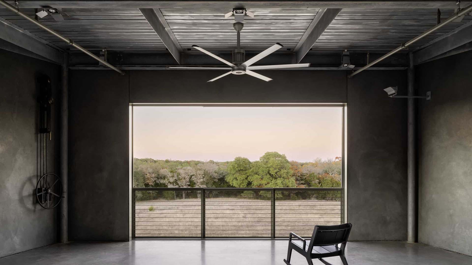 A minimalist room with a large ceiling fan, an empty black chair, and an open wall framing a view of trees and a fence outside.
