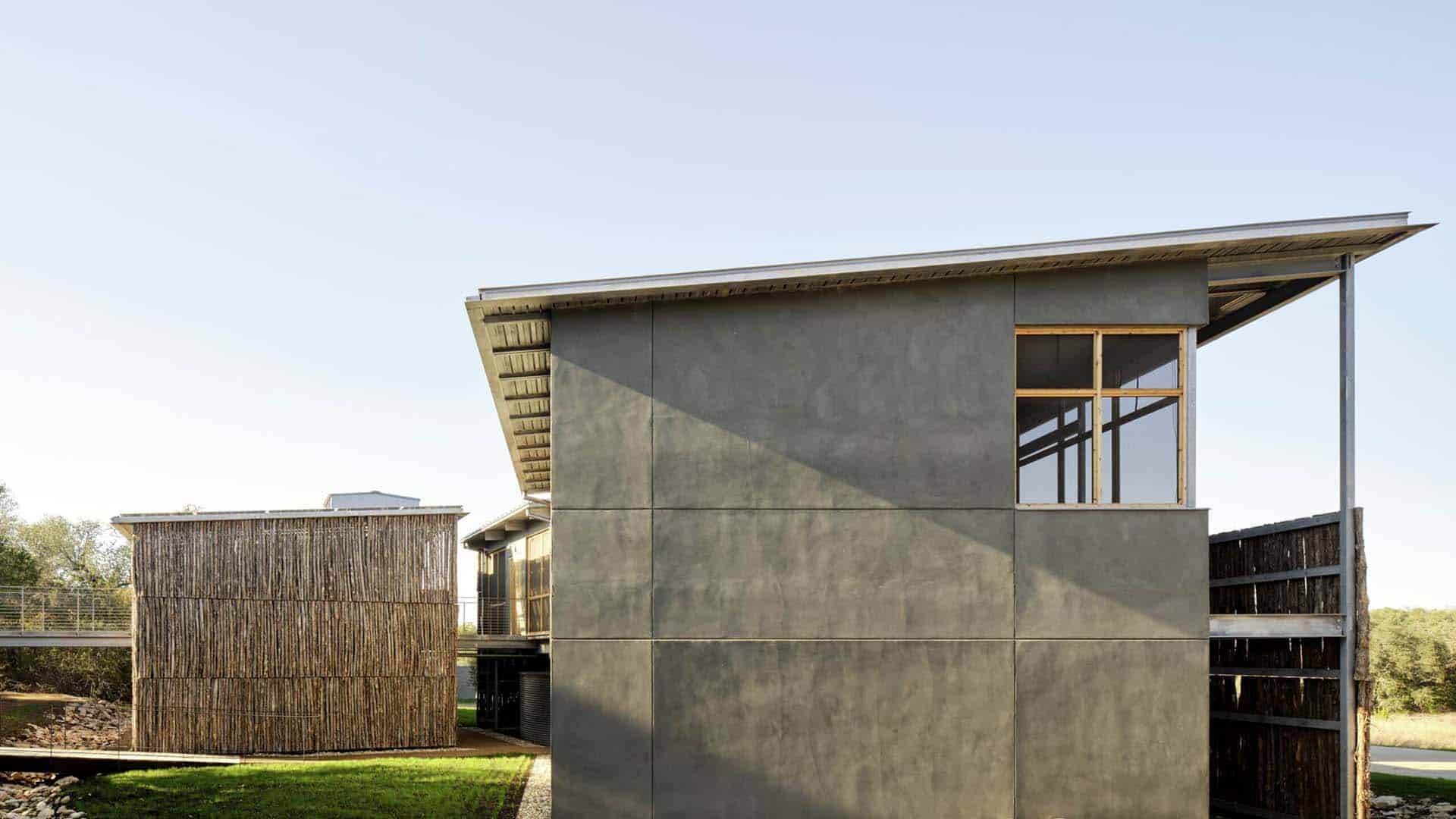 Modern building with a slanted metal roof and large windows, next to a cylindrical structure with vertical wooden slats, under a clear blue sky.