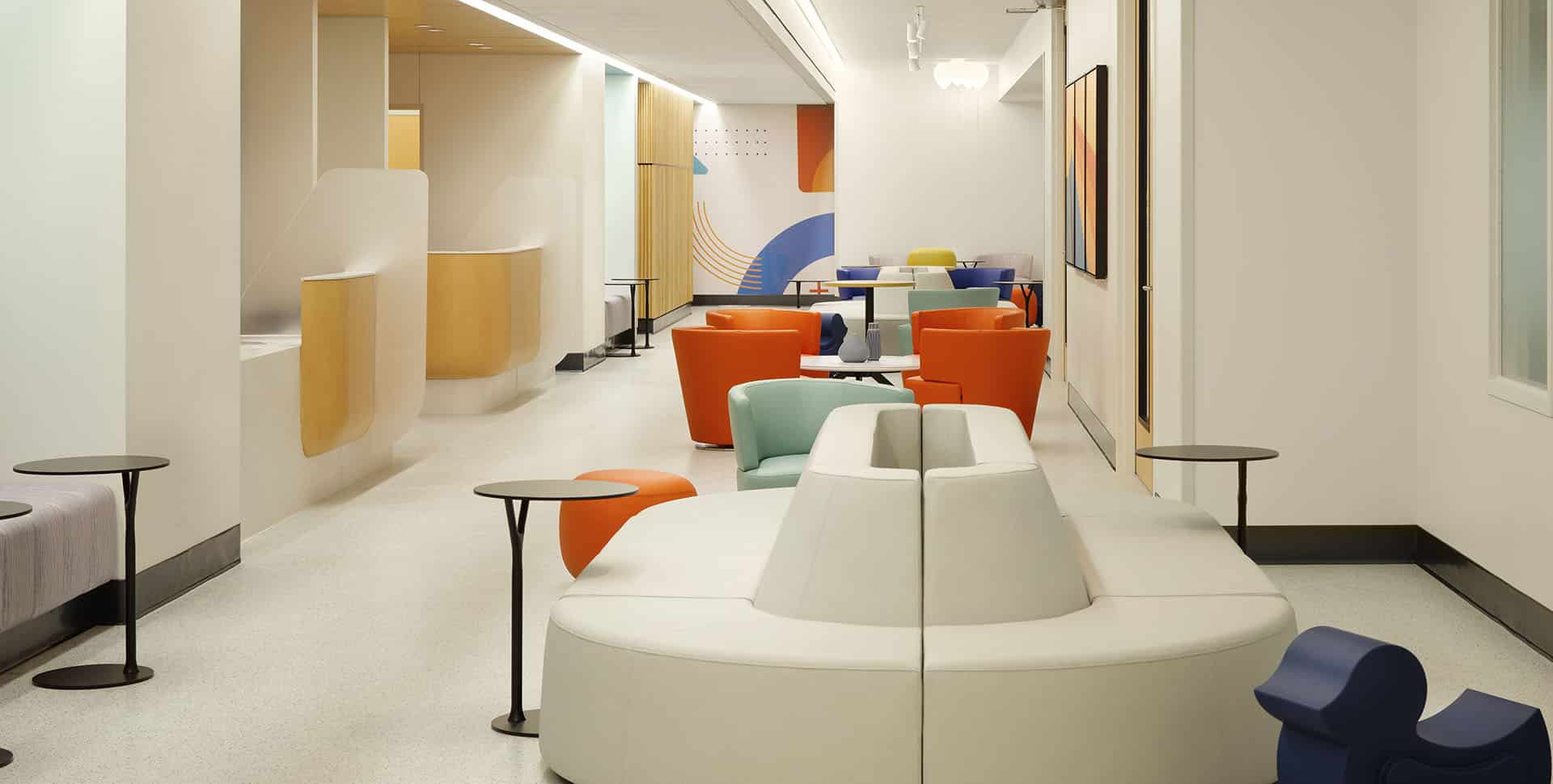 Modern waiting area with colorful chairs, small tables, and a white curved sofa, set in a bright space with neutral walls and abstract art.