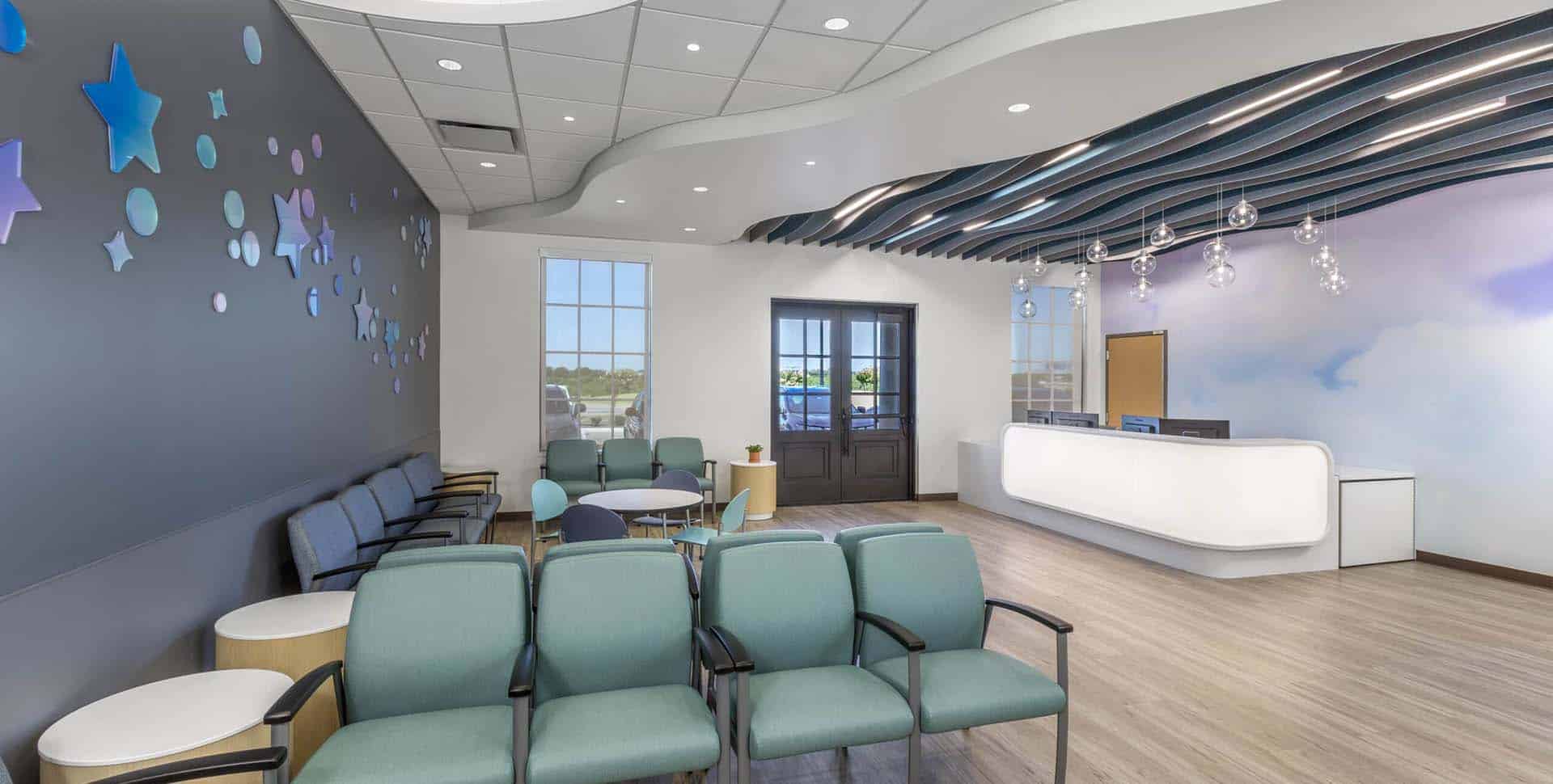 Modern waiting room with teal chairs, a curved reception desk, wood flooring, and star decorations on the wall, with natural light coming in through windows.