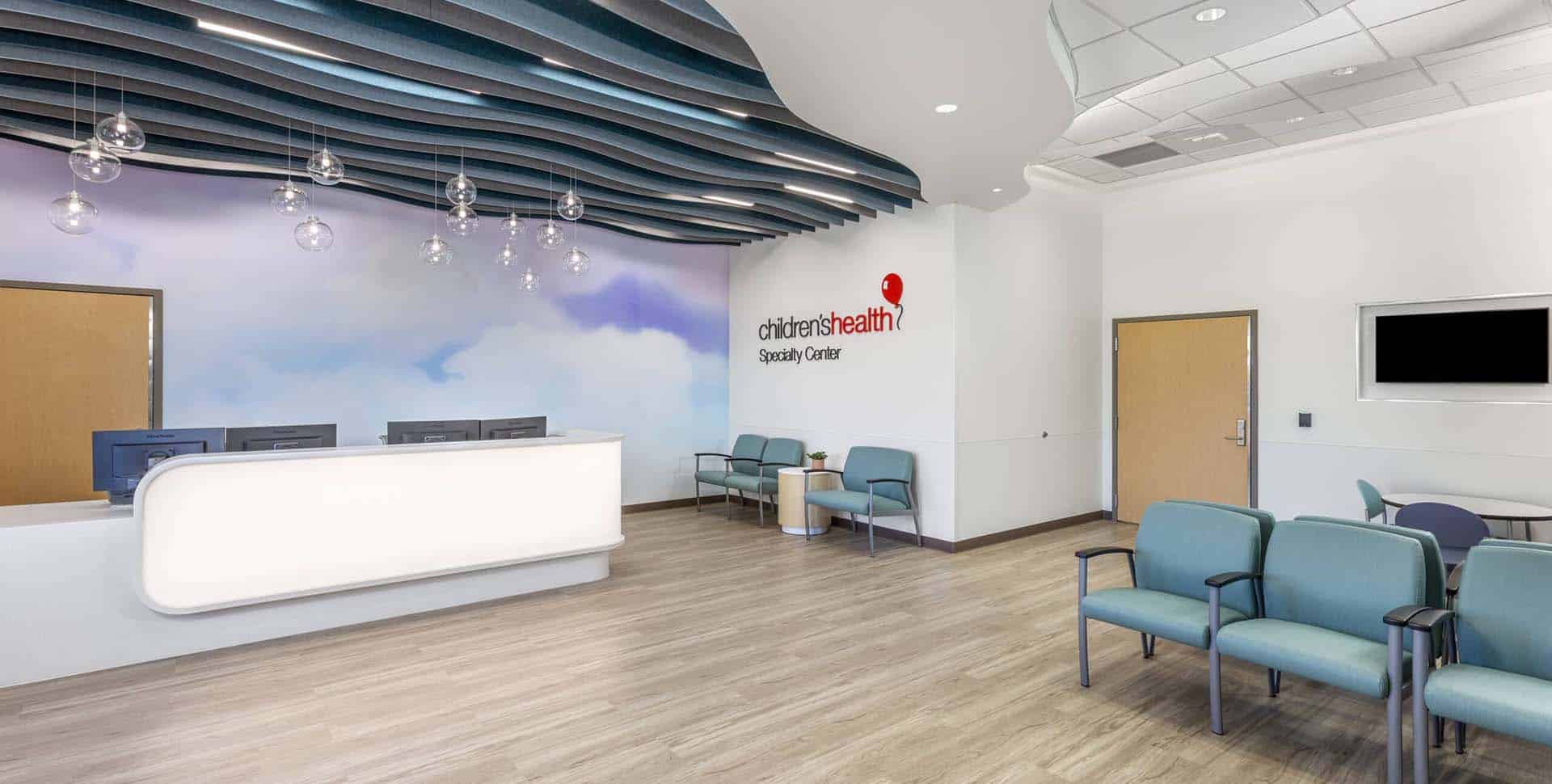 Modern medical waiting area with teal chairs, a reception desk, and overhead light fixtures; "children's health Specialty Center" sign on the wall.