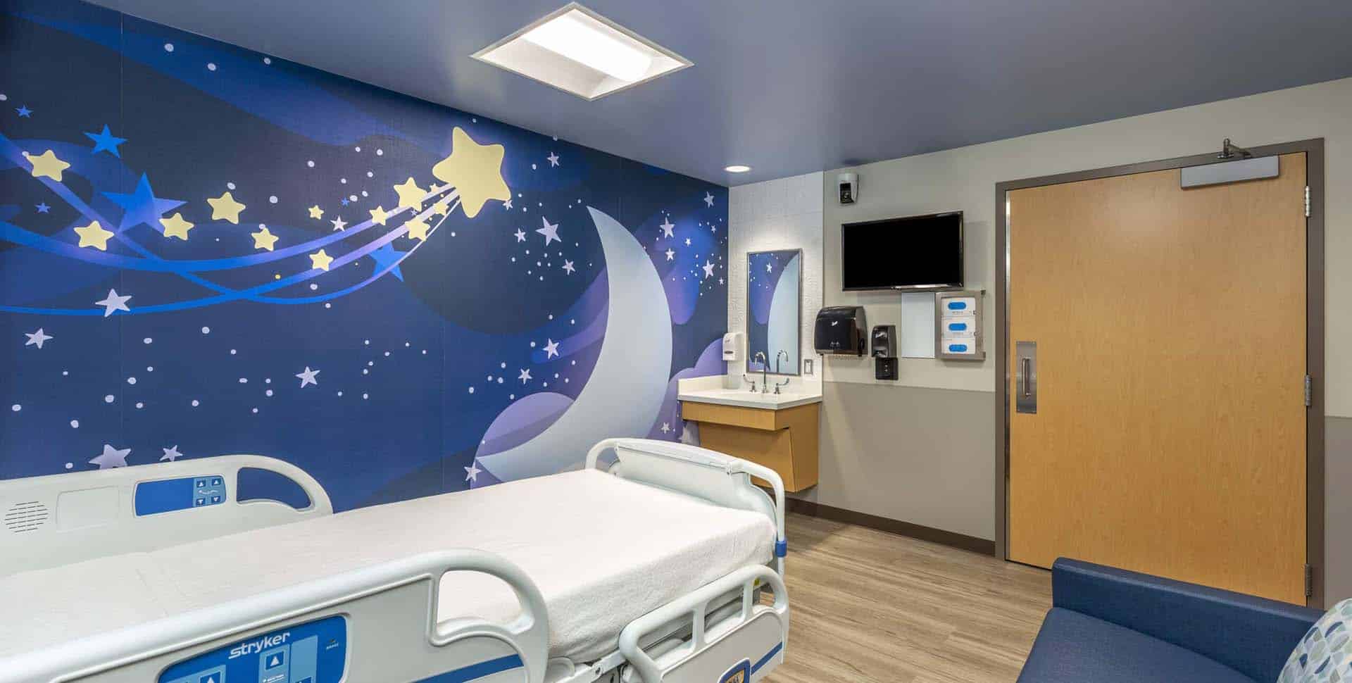 A hospital room with a children’s bed, a wall featuring a moon and stars mural, a TV, a sink, soap dispensers, and a wooden door.