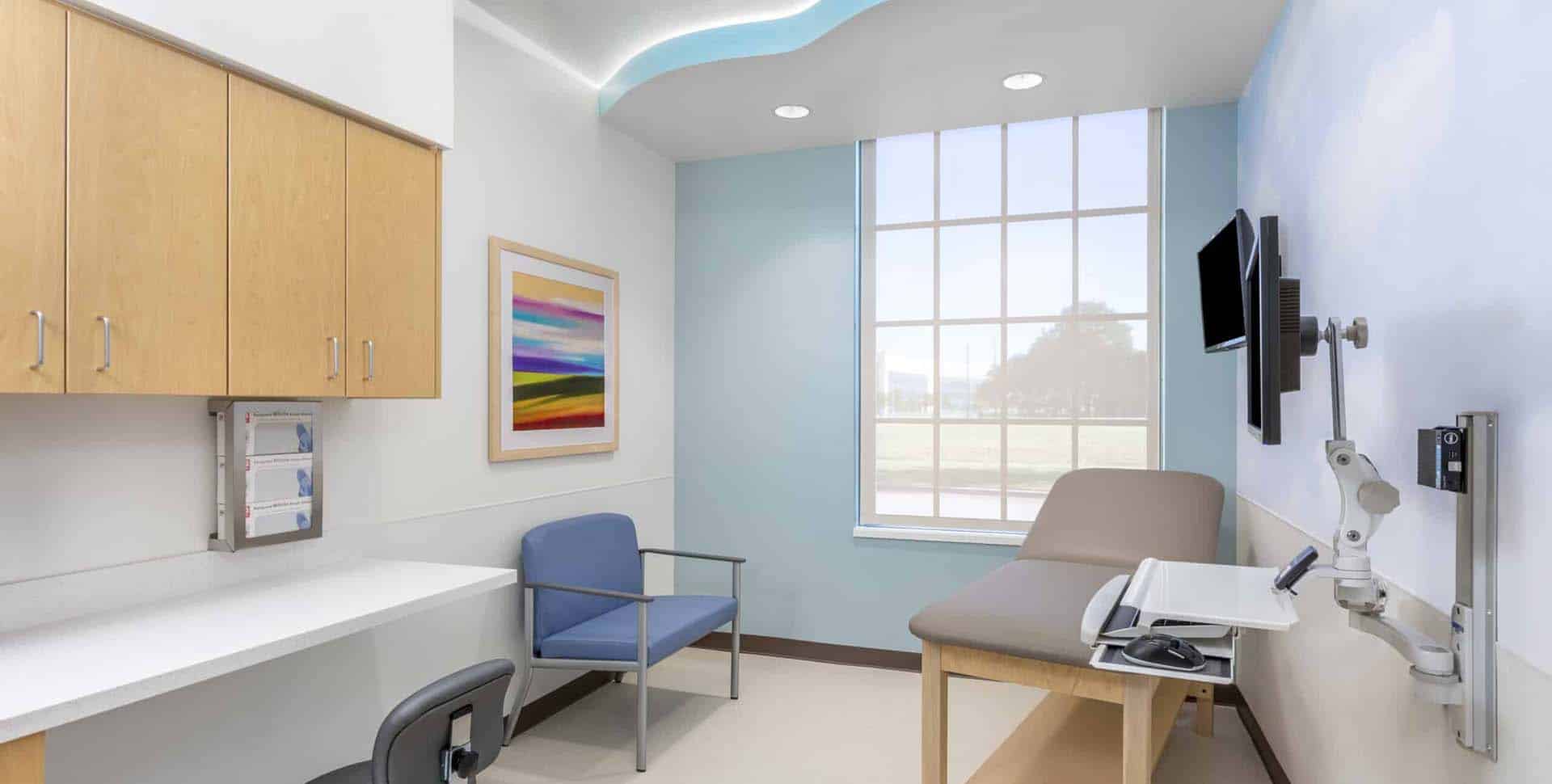 A clean, modern medical exam room with a desk, chairs, exam table, wall-mounted monitor, cabinets, and a large window letting in natural light.