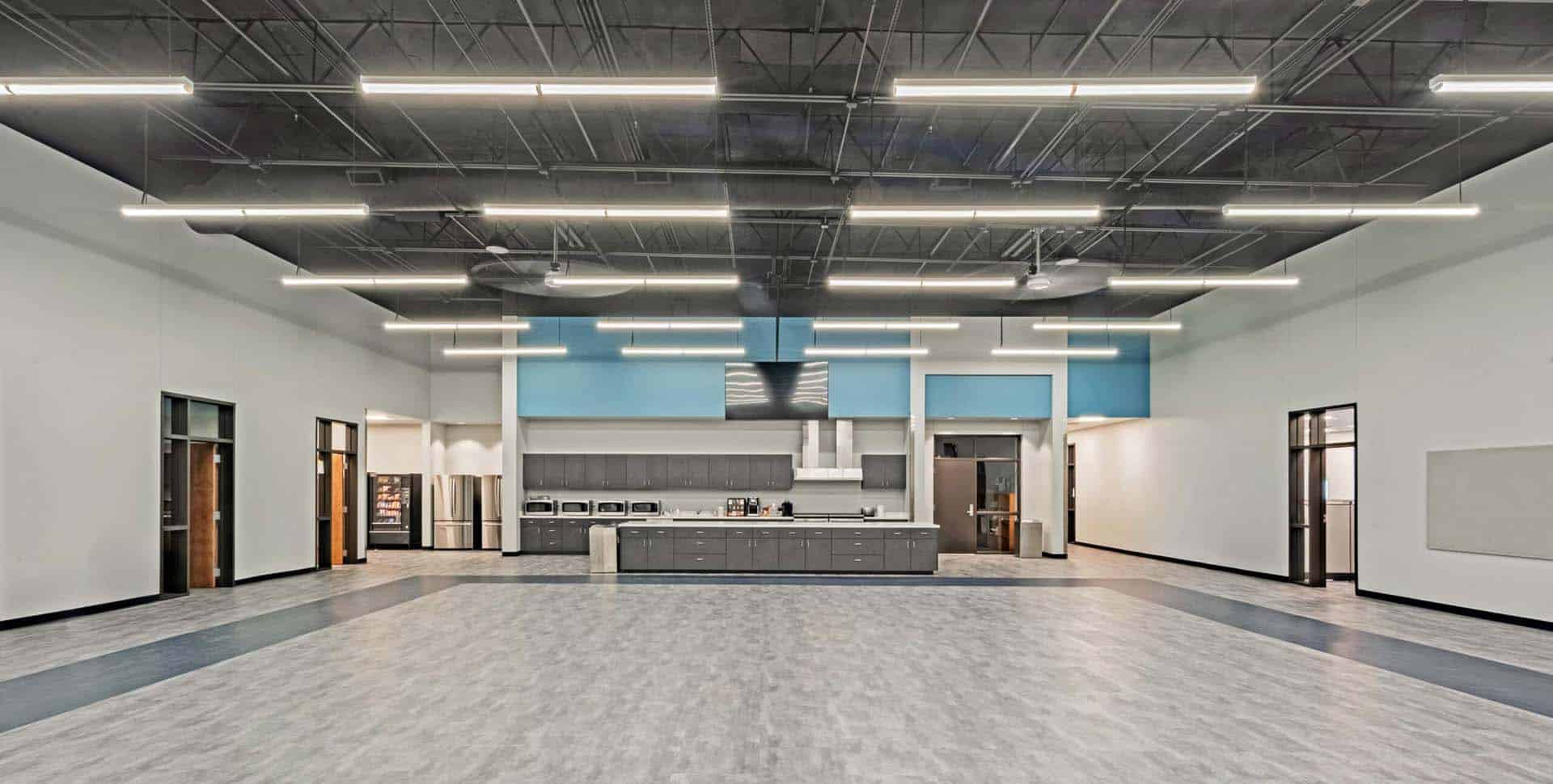Spacious, modern multipurpose room with a kitchenette, gray flooring, blue and white walls, and overhead fluorescent lighting.
