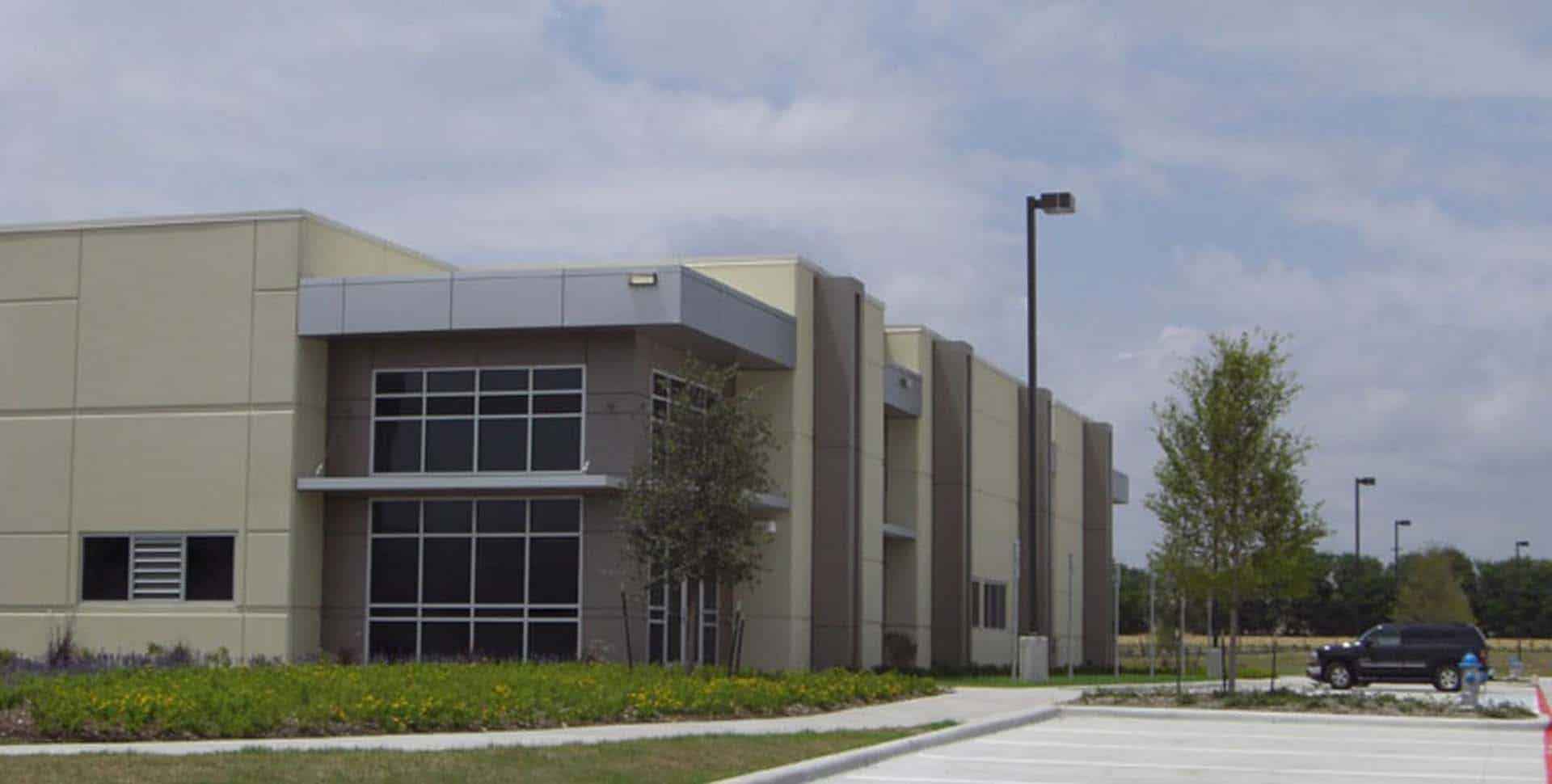 A modern, beige commercial building with large windows, surrounded by landscaping and an empty parking lot under a partly cloudy sky.