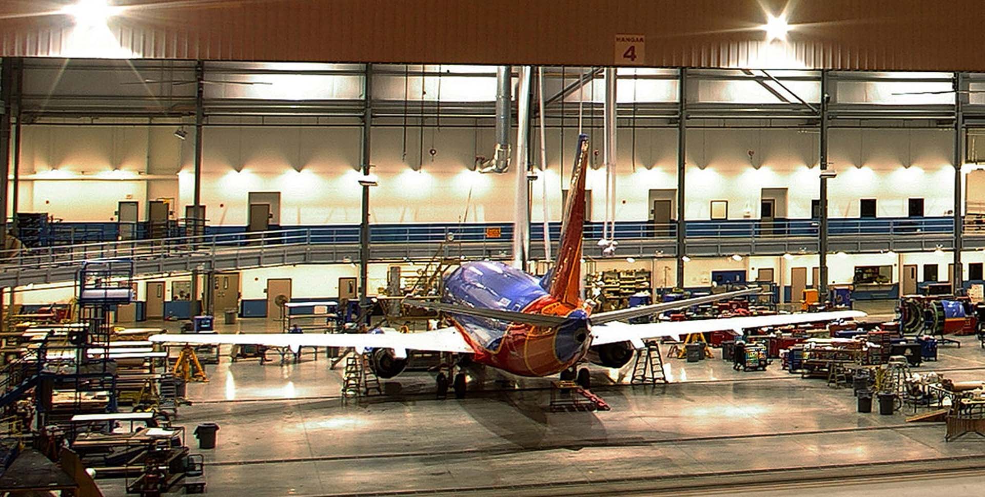 A commercial airplane is parked inside a brightly lit maintenance hangar, surrounded by equipment and workstations.