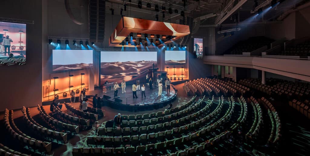 A large auditorium with tiered seating, a central stage, and multiple performers; desert landscape visuals are displayed on large screens behind the stage.