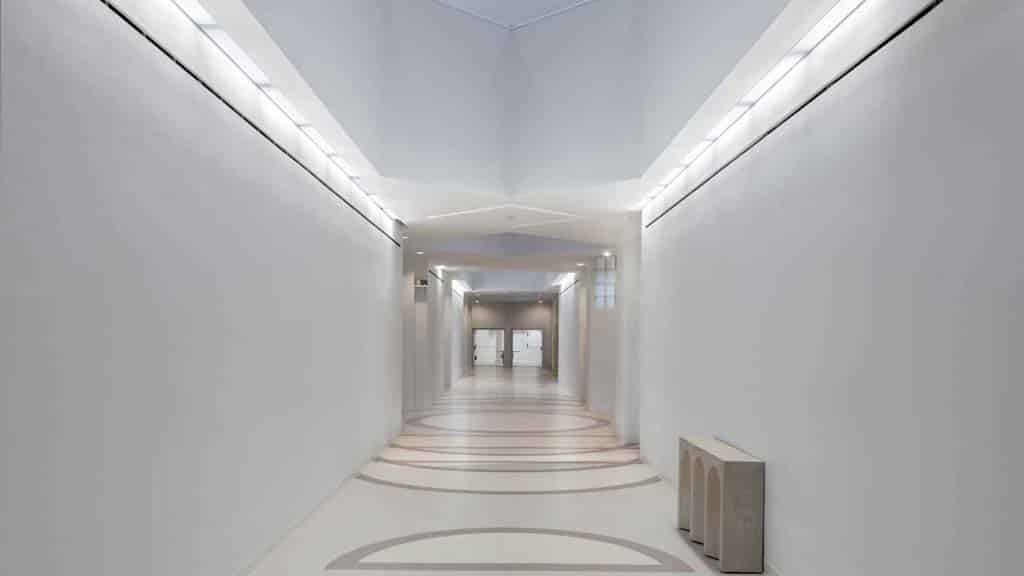 A modern, empty hallway with white walls, geometric ceiling lights, and decorative floor patterns leads to glass doors at the far end.