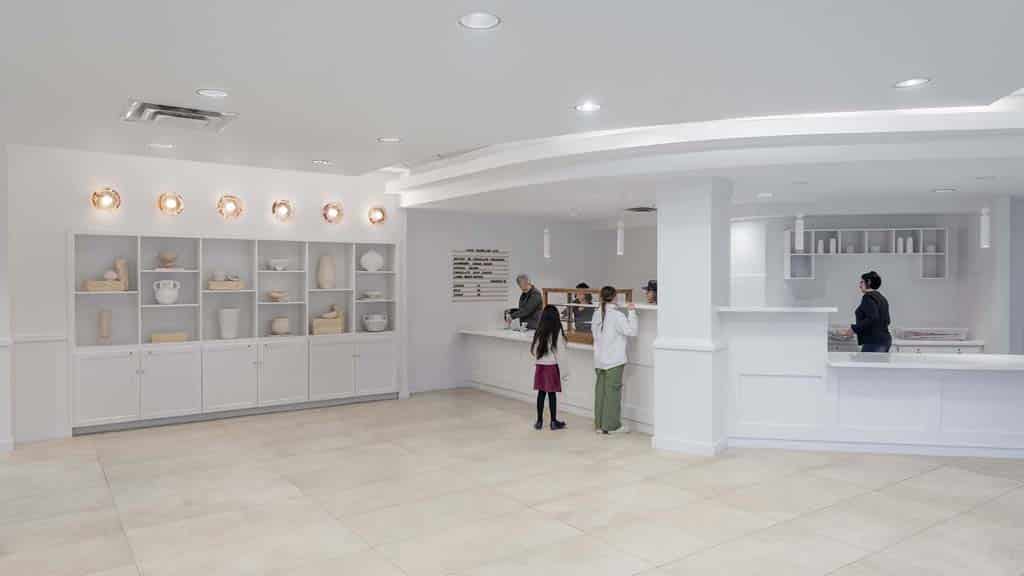 A modern, white bakery interior with display shelves on the left and several people interacting at the counter on the right.