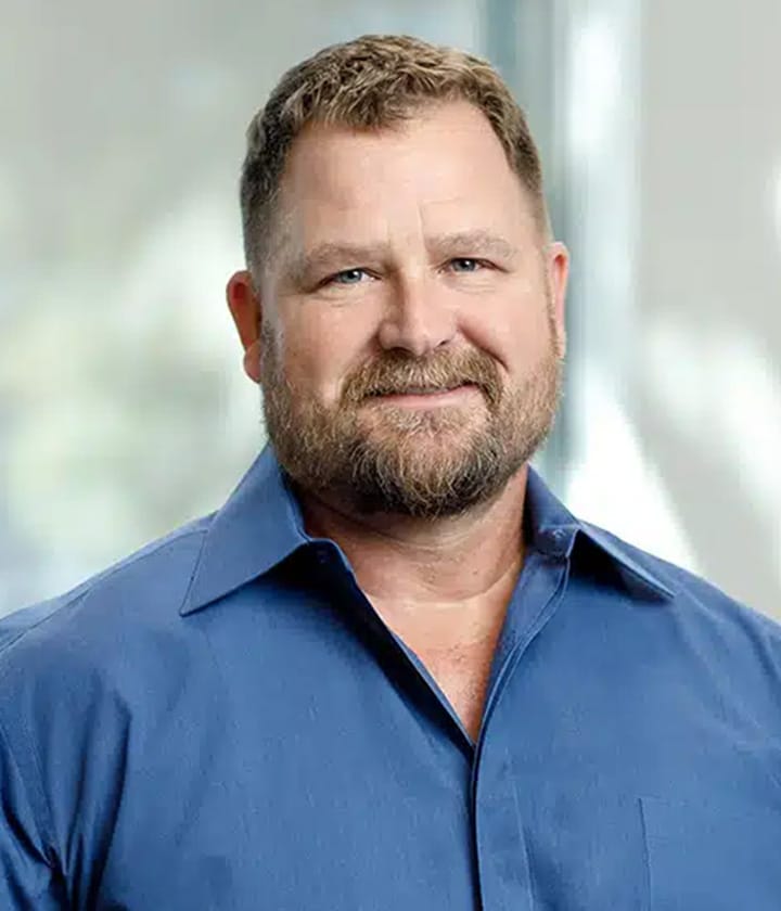 A man with short hair and a beard wearing a blue collared shirt, standing indoors in front of a softly blurred background.