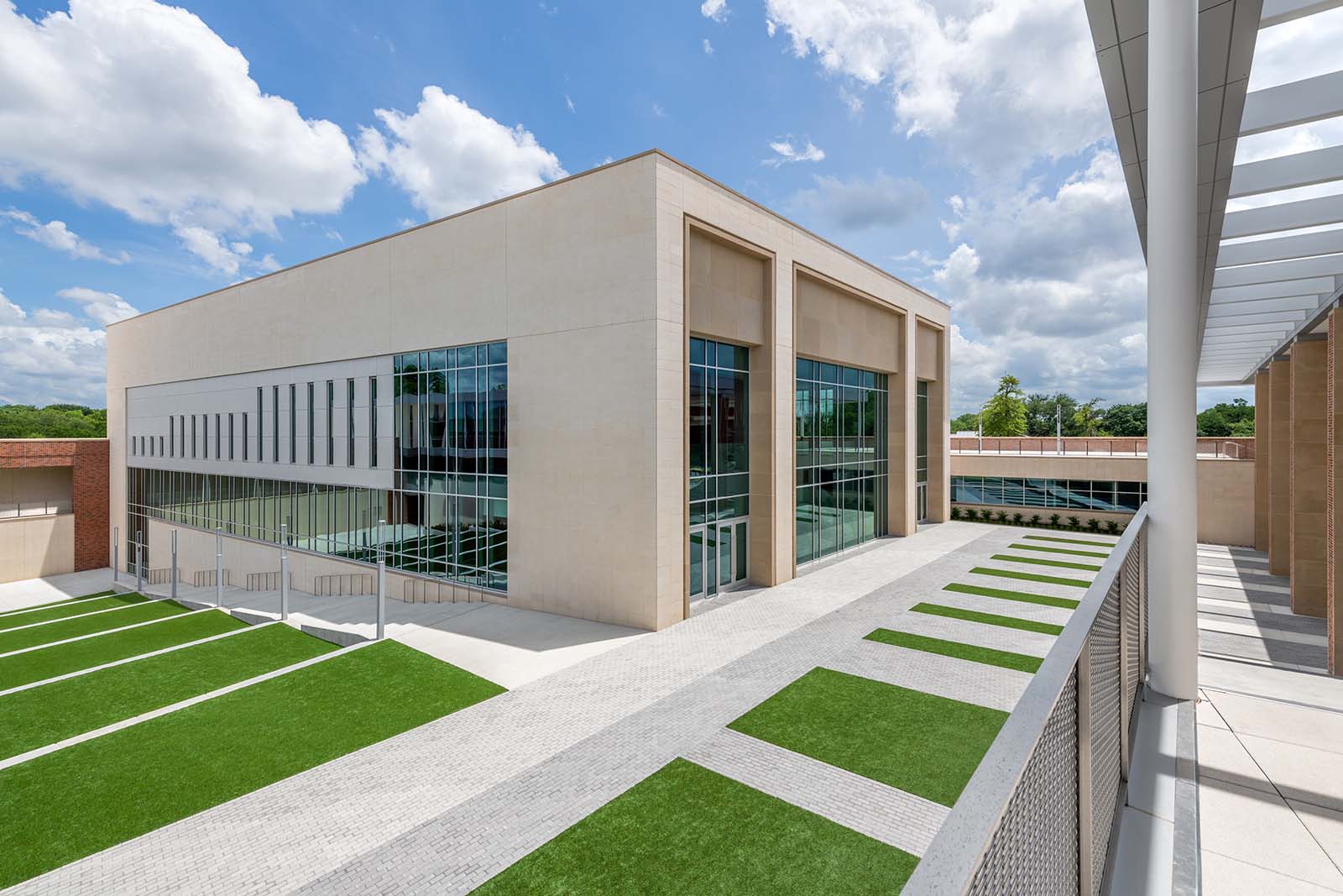 Modern two-story building with large glass windows and beige walls, surrounded by neatly manicured green lawns and a paved walkway under a partly cloudy sky.