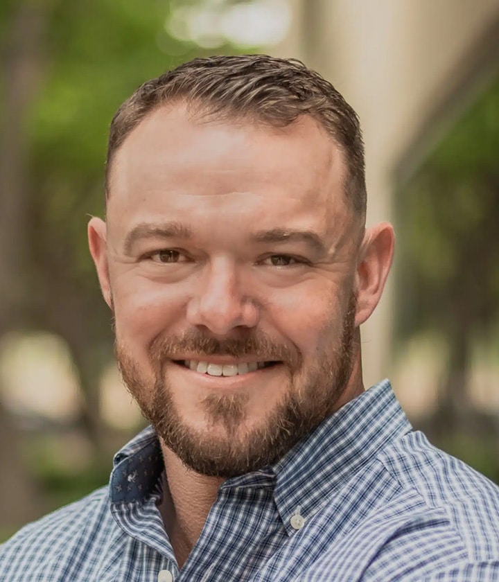 A man with short brown hair and a trimmed beard, wearing a blue checked shirt, smiling outdoors with a blurred background.