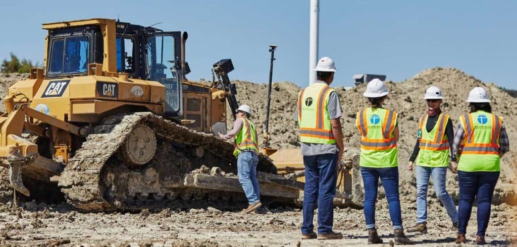 Five construction workers wearing safety vests and helmets stand near a large CAT bulldozer at a dirt construction site under a clear sky.