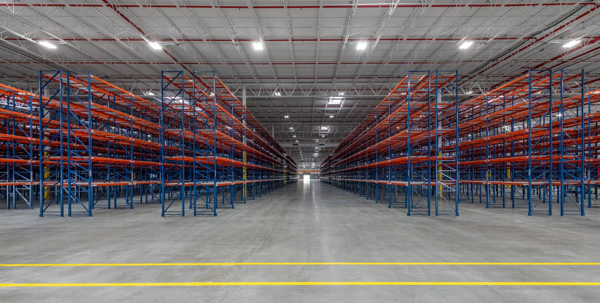 Large empty warehouse with rows of tall, blue and orange metal storage racks on either side of a wide central aisle under bright overhead lighting.