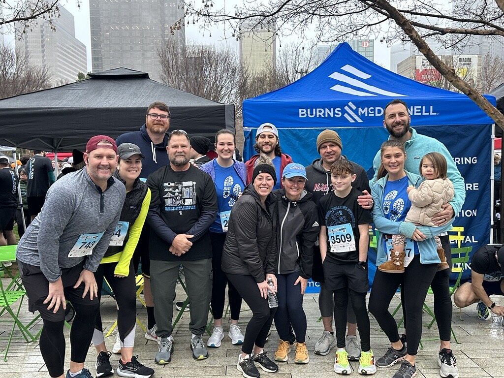 A group of people wearing athletic clothing and race bibs pose together in front of a blue Burns & McDonnell tent at an outdoor event.