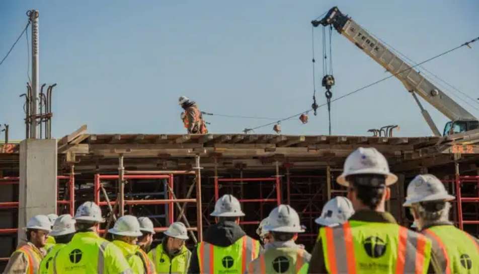 Construction workers in safety vests and helmets observe a team member on scaffolding, while a crane operates in the background at a building site.