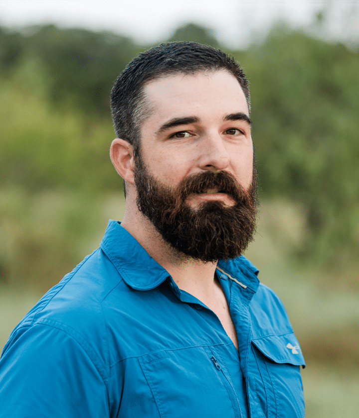 A man with a dark beard and short hair wearing a blue collared shirt stands outdoors with greenery in the background.