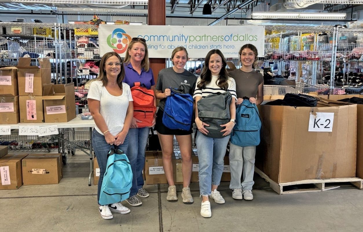 Five people stand in a warehouse, each holding a backpack, in front of a sign that reads "Community Partners of Dallas" and boxes labeled by grade levels.
