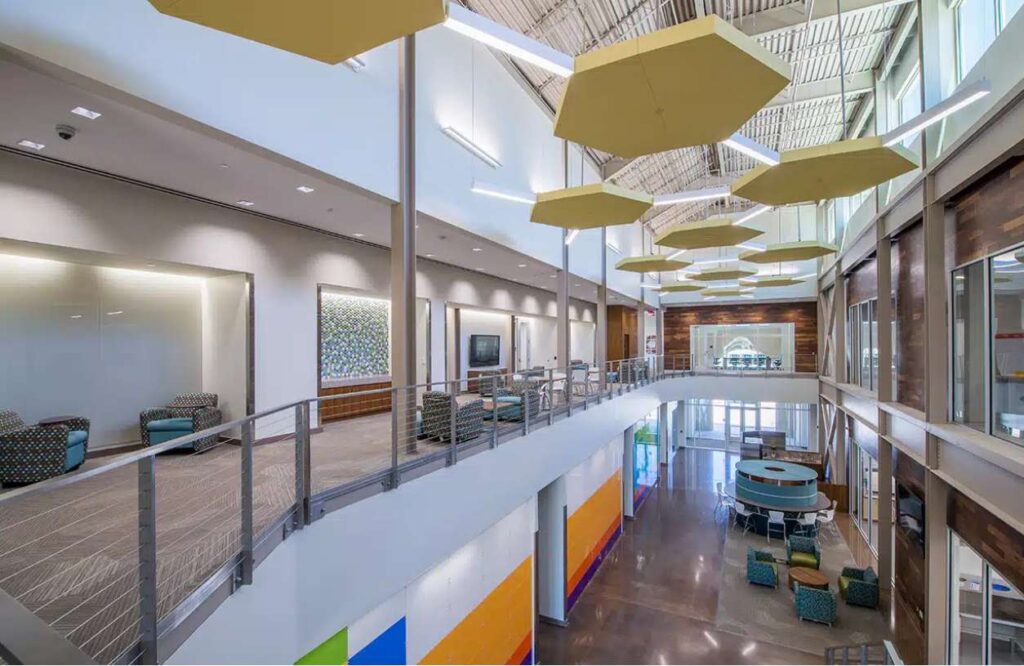 A modern, two-story interior space with large windows, bright geometric wall accents, hanging yellow ceiling panels, and a mezzanine with lounge seating areas.