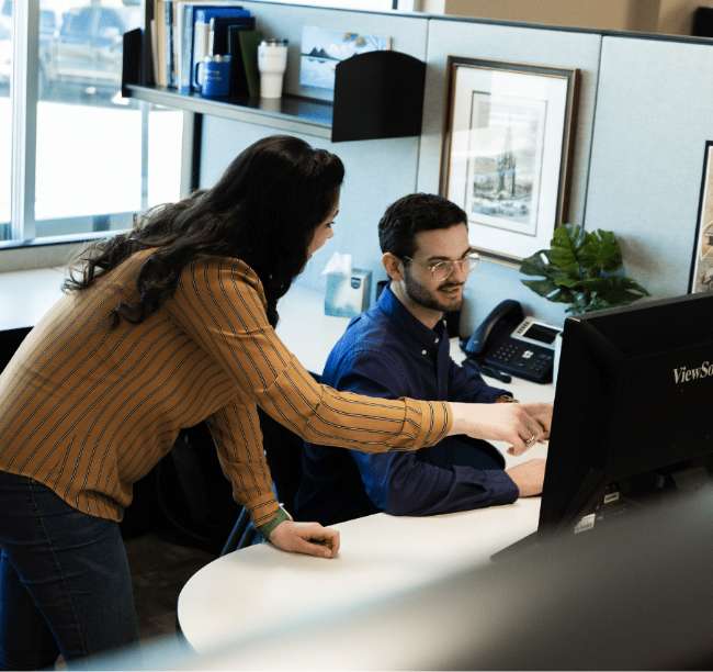 A woman stands beside a seated man at an office desk, pointing at his computer screen while they discuss something.