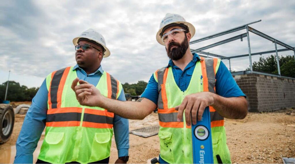 Two construction workers wearing safety vests and helmets stand on a construction site; one gestures while holding a level tool.