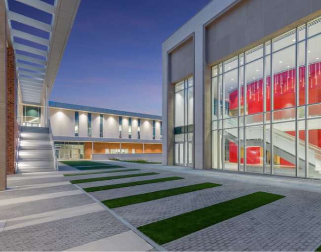 Modern building courtyard at dusk with glass walls, red interior accents, paved walkway, green grass strips, and exterior staircases.