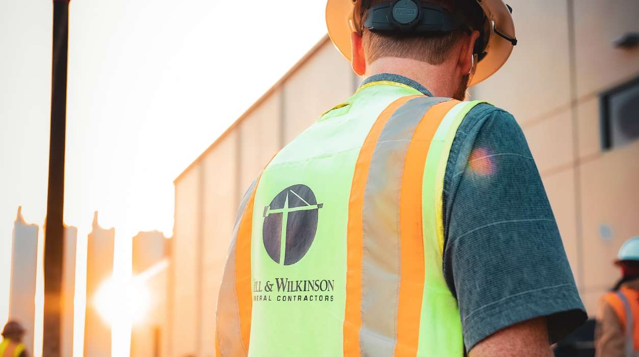 A construction worker wearing a hard hat and a high-visibility vest with the JL & Wilkinson Metal Contractors logo stands outdoors near a building at sunset.